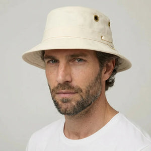 A bearded man in a white shirt wears the TILLEY - ICONIC T1 BUCKET HAT IN NATURAL with UPF 50+ protection and metal eyelets, looking directly at the camera against a plain background.