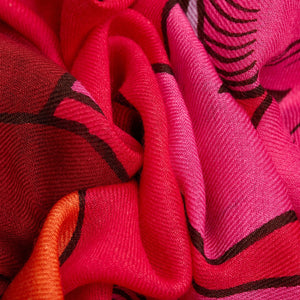 Close-up of the INOUI EDITIONS - CAVALE WOOL SCARF IN RED by INOUI, featuring crumpled wool in red, pink, and orange with dark brown abstract lines. Its woven texture and bold style make it a perfect winter accessory.
