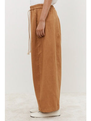 A person in MEIMEIJ Linen Drawstring Pants, featuring a relaxed wide-leg brown silhouette with a white drawstring, pairs them with white shoes against a plain light background, showcasing their adjustable fit for a casual look.