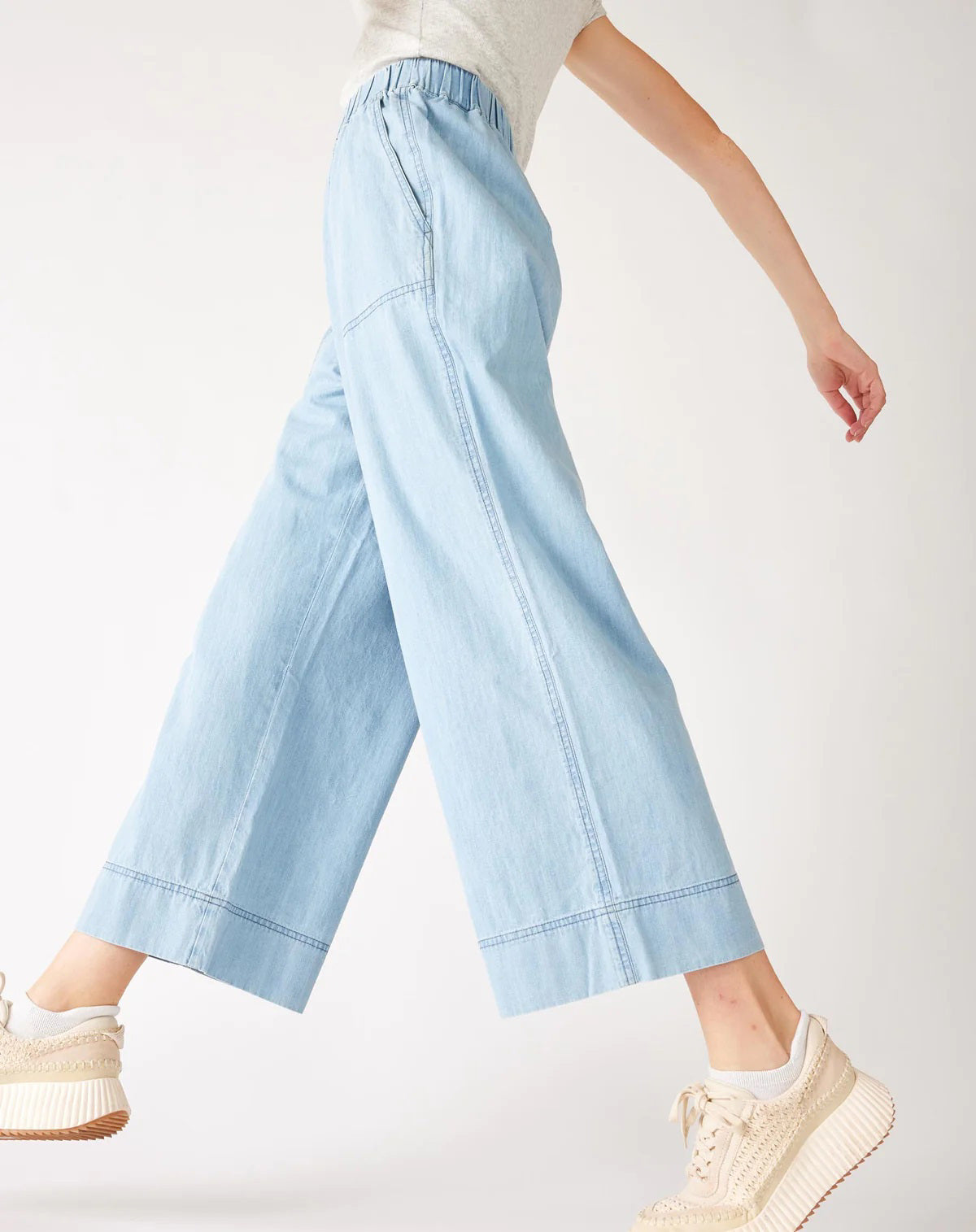 Wearing MERSEA - SAMMIE WIDE LEG PANT in light blue chambray with a pull-on waistband and front patch pockets, styled with a light top and cream sneakers, the person stands against a plain background.