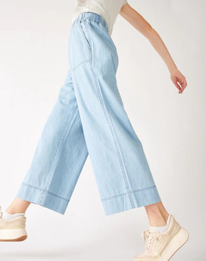 Wearing MERSEA - SAMMIE WIDE LEG PANT in light blue chambray with a pull-on waistband and front patch pockets, styled with a light top and cream sneakers, the person stands against a plain background.