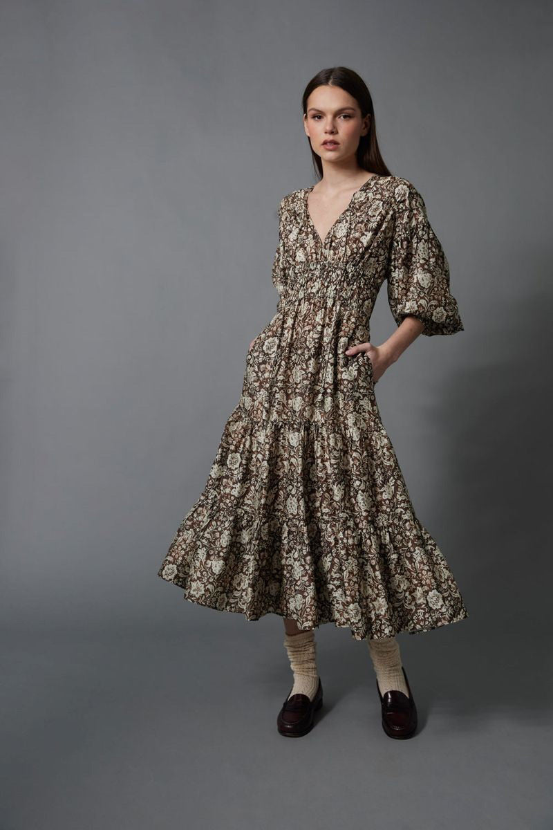 A woman stands against a gray background wearing the HOPE FOR FLOWERS TIERED PEASANT DRESS with puffed sleeves and a shirred bodice. She has her hands in its pockets and pairs dark loafers with light-colored socks.