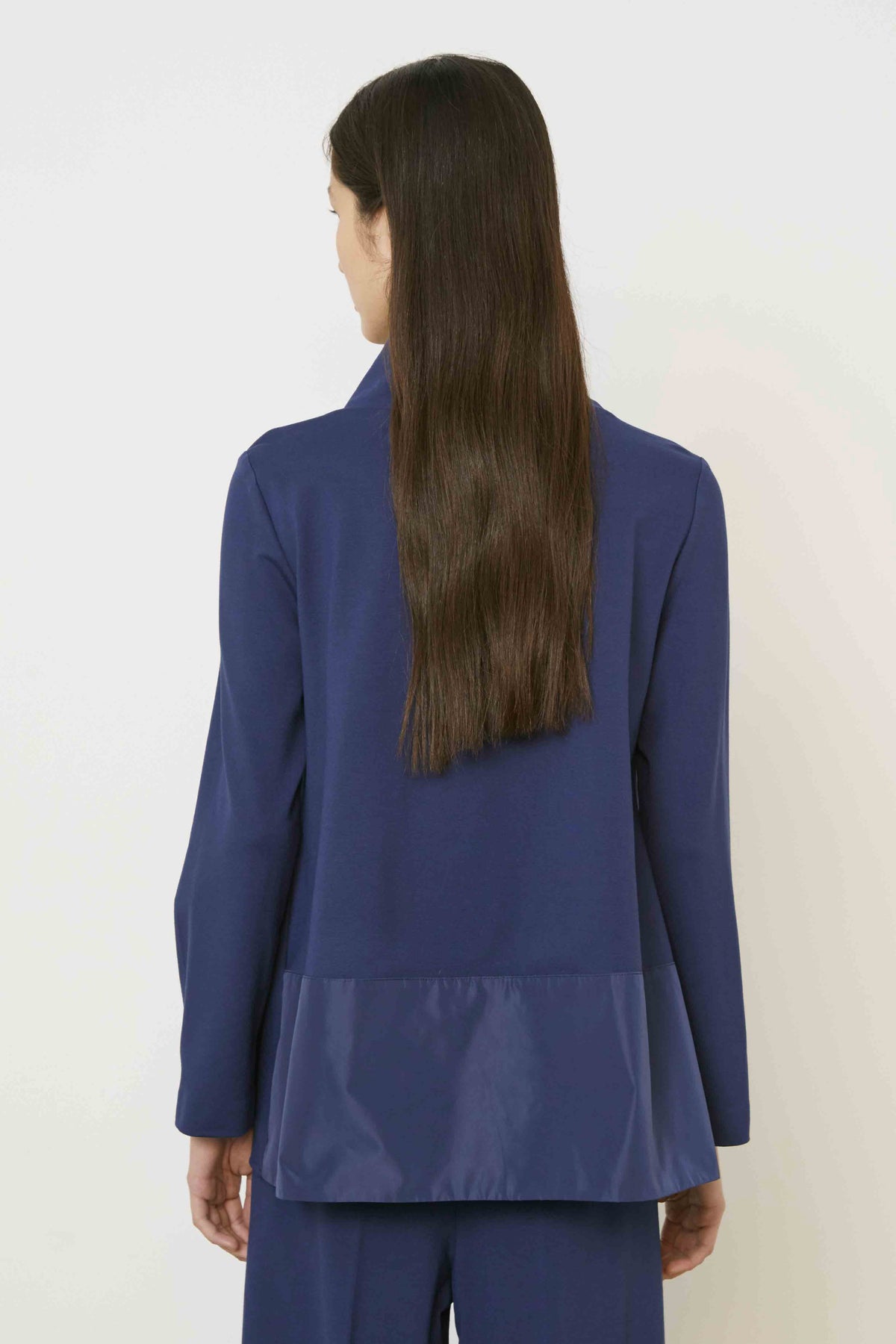 A woman with long brown hair, seen from behind, wears the MEIMEIJ Mixed Media Cowl Neck Top in dark blue with a shiny panel at the bottom and matching pants. She stands against a plain, light background.