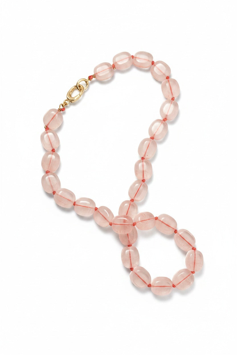 The SANDRINE FRANCE STUDIO - STAFNIE NECKLACE IN ROSE CORAL by SANDRINE FRANCE STUDIO features oval-shaped, translucent pink glass beads artfully strung together with a gold-plated clasp. Delicate red knots separate each bead, all elegantly arranged against a white background.