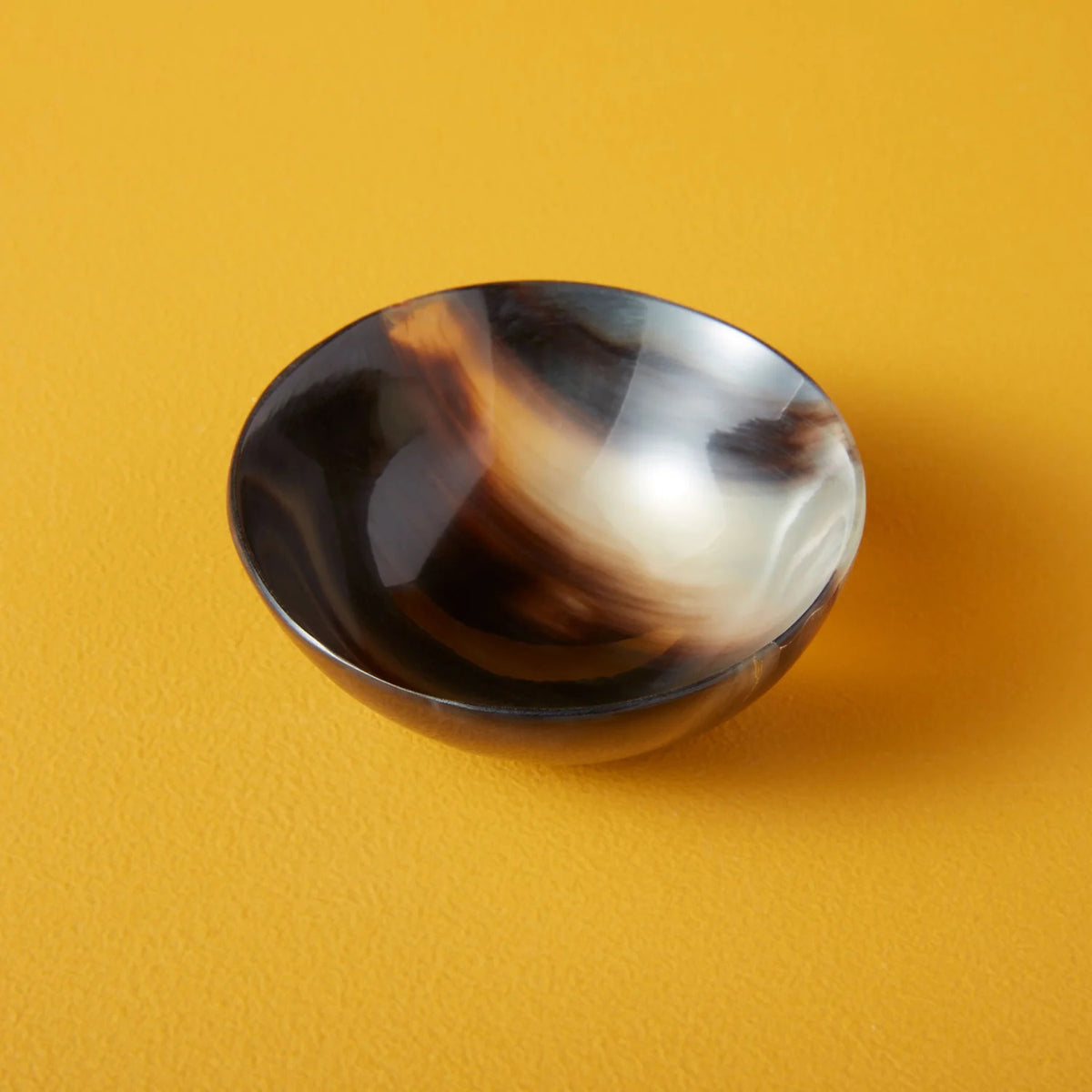 The SPLATTER HORN BOWL SMALL from BE HOME, INC is a small, round bowl made from water buffalo horn, showcasing an intricate marbled pattern in shades of brown, black, and white. Ethically crafted and handmade in Vietnam, the bowl boasts a polished, glossy finish and is set against a vibrant yellow surface.