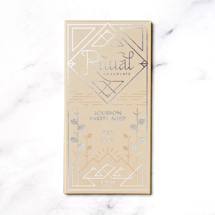 The image shows a light brown rectangular package of Ritual Chocolate labeled &quot;RITUAL CHOCOLATE - BOURBON BARREL AGED CHOCOLATE BAR 75%.&quot; This limited edition chocolate, crafted from aged cacao nibs, features geometric designs in silver and white and contains 2.12 oz (60g) of chocolate. The background is a marble surface.