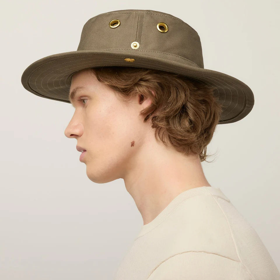 A young person with light skin and wavy brown hair wears a beige TILLEY - CLASSIC T3 HAT from TILLEY, featuring gold accents, paired with a beige shirt and looking at the camera against a plain light background.
