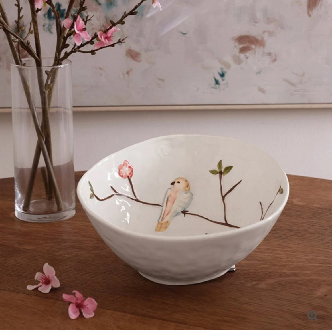 The BEATRIZ BALL - CERAMIC BIRD ON BRANCH BOWL by BEATRIZ BALL rests on a wooden table beside a glass vase with pink flowering branches.