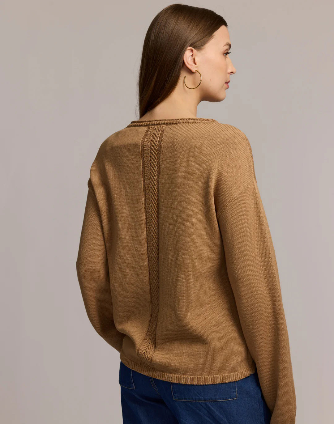 A woman with long brown hair and large hoop earrings models the HINSON WU AUDREY Long Sleeve Cotton Silk Sweater in tan with blue jeans, standing against a plain beige background, one hand raised near her shoulder.