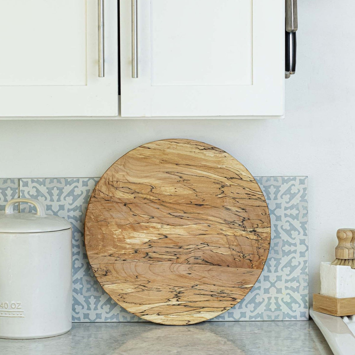 A round wooden cutting board with natural grain rests upright on a kitchen counter, perfect paired with the PETERMANS BOARDS &amp; BOWLS INC SPENCER PETERMAN - SPALTED LAZY SUSAN 20" for extra artisan charm.