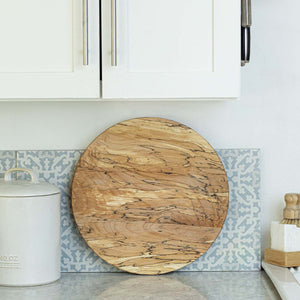 A round wooden cutting board with natural grain rests upright on a kitchen counter, perfect paired with the PETERMANS BOARDS & BOWLS INC SPENCER PETERMAN - SPALTED LAZY SUSAN 20" for extra artisan charm.