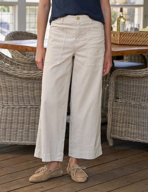 A person wearing the FRANK & EILEEN Limerick Cropped Wide Leg Carpenter Pant in Italian Performance Linen Cement, styled with tan lace-up shoes, stands indoors on a wooden floor with wicker furniture in the background.