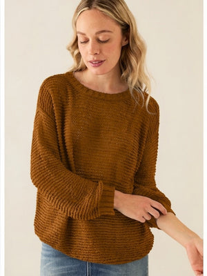 A woman with wavy blonde hair wears a BASE MILANO - CREWNECK COTTON BLEND SWEATER in a relaxed fit and blue jeans, standing against a plain light background as she looks down and gently pulls up one sleeve.