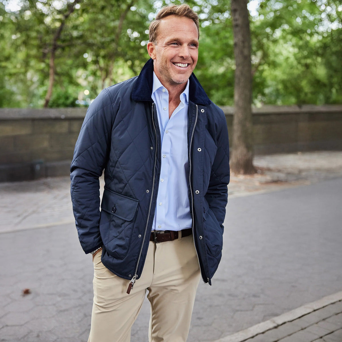 A man with short blond hair wears a navy quilted jacket, light blue shirt, and COLLARS AND CO Apex Tech Pants in Light Beige as he sits on a green bench outdoors, looking at the camera with his hands clasped.