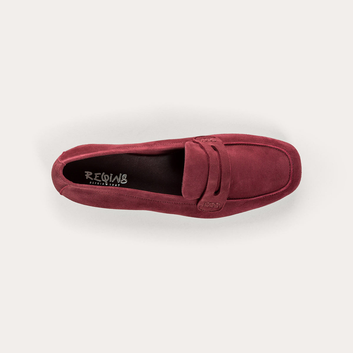 Close-up of the REQINS - HEMA SUEDE LOAFER IN BORDEAUX by REQINS, showcasing artisanal craftsmanship with burgundy suede, detailed stitching, a front strap, and a soft leather insole, displayed on a light surface.