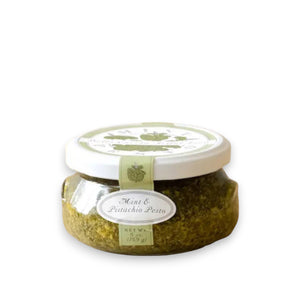 A small glass jar filled with green pesto labeled "BELLA CUCINA - MINT PISTACHIO PESTO" is shown. The white lid has a green design, and a seal runs from the lid to the base, marked with the same label. Made with olive oil and fresh mint leaves, the jar sits on a reflective surface against a plain background.