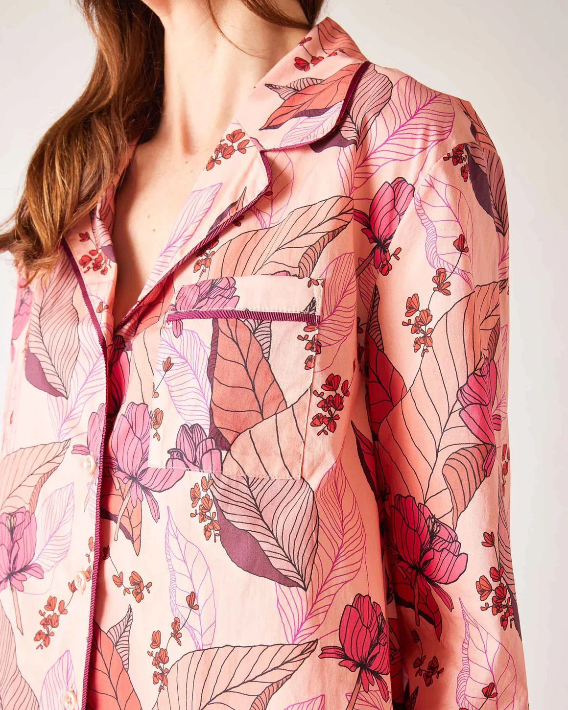 A woman stands barefoot wearing the MERSEA Over The Moon PJ Set in Rosewood by MERSEA—light pink cotton pajamas with vibrant pink and purple florals, a button-up shirt, wide-leg pants, and a comfy stretch waistband. Her face is partially out of frame.