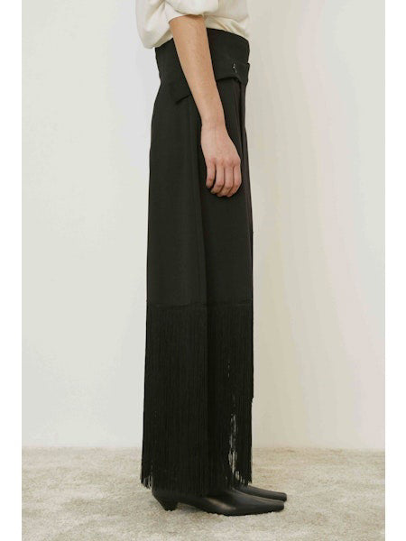 A person stands against a plain background wearing the MEIMEIJ - FRINGE WRAP SKIRT with an asymmetric hem, paired with a light top and black boots.