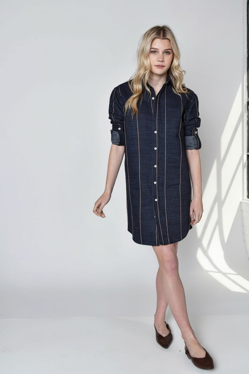 A person with long blonde hair wears the EMILY PHILLIPS Denim and Brown Stripe Girlfriend Mini Dress, styled with brown flats and rolled sleeves, standing against a plain light background.