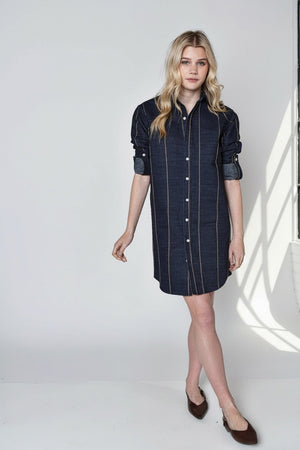 A person with long blonde hair wears the EMILY PHILLIPS Denim and Brown Stripe Girlfriend Mini Dress, styled with brown flats and rolled sleeves, standing against a plain light background.