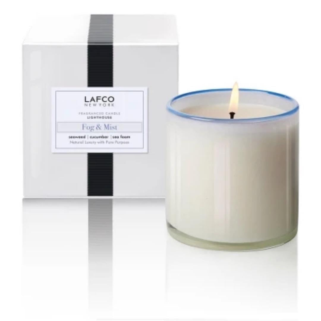 A LAFCO - FOG MIST, LIGHTHOUSE scented candle, hand-poured into a clear glass container with a lit wick, sits next to its elegant white box packaging. The box features black vertical stripes and a label that reads &quot;LAFCO New York&quot; and &quot;Fog &amp; Mist.&quot; Luxurious candles like these add sophistication to any room.