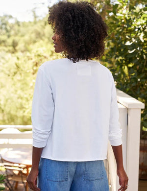 A woman with curly hair smiles outdoors, wearing the FRANK & EILEEN Ella Long Sleeve Boatneck Tee in Heritage Jersey White from the California Heritage Collection, and displaying a ring on her left hand.