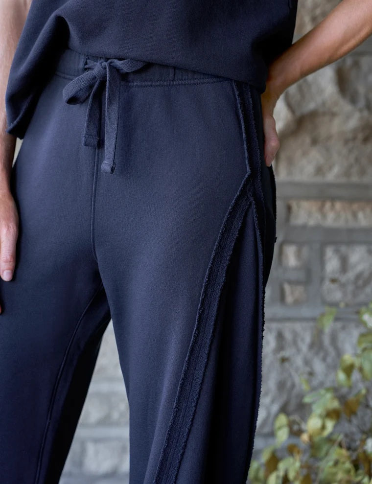 A woman with wavy brown hair smiles as she stands in a bright room, wearing the FRANK & EILEEN Bibi Barrel Leg Sweatpant in Classic Navy paired with a matching short-sleeve top and tan loafers.