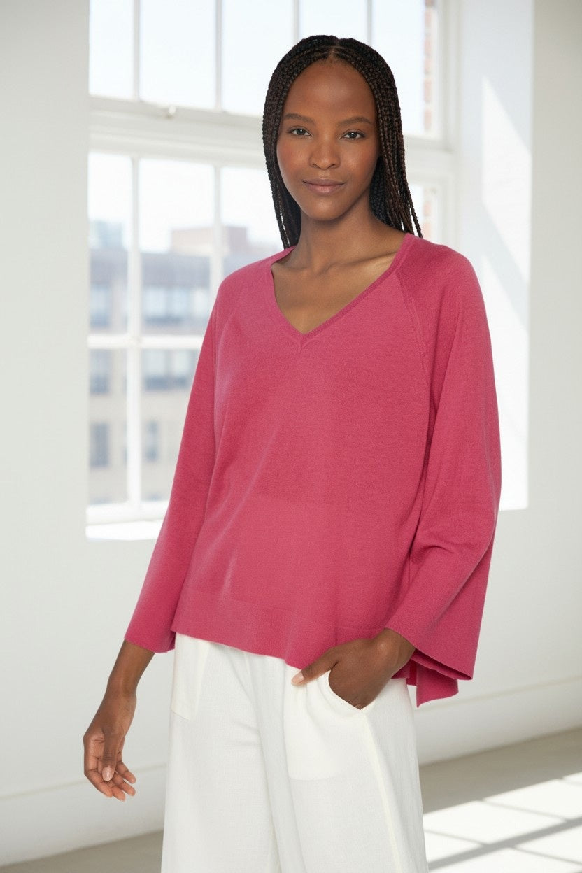 A discerning dresser with long braided hair wears the KINROSS CASHMERE EASY HI LOW VEE NECK SWEATER in orange and white pants. They stand against a plain background, smiling softly, with one hand in their pocket and the other by their side.