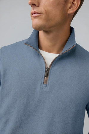 A man with short light brown hair stands in profile, wearing the MACK WELDON Ace 1/2 Zip Sweater in Herringbone and dark pants, one hand in his pocket, against a plain light gray background.