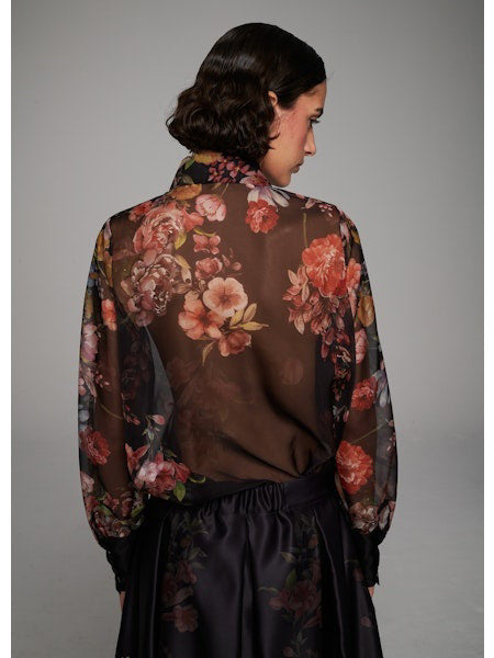 A woman with wavy dark hair models the MILVA MI - FLORAL VOILE BLOUSE by MILVA—a semi-sheer, long-sleeve top with a dark floral print and black collar—paired with a high-waisted skirt, standing before a plain gray background.
