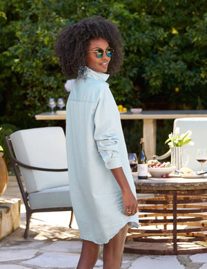 The FRANK AND EILEEN - MARY CLASSIC SHIRTDRESS in Classic Blue Tattered Wash by FRANK & EILEEN is a light blue, long linen shirtdress featuring a button-down front and long sleeves rolled up to three-quarter length. It has a single chest pocket and resembles the casual elegance of Famous Denim with its textured fabric and slightly frayed hem, displayed against a textured white background.