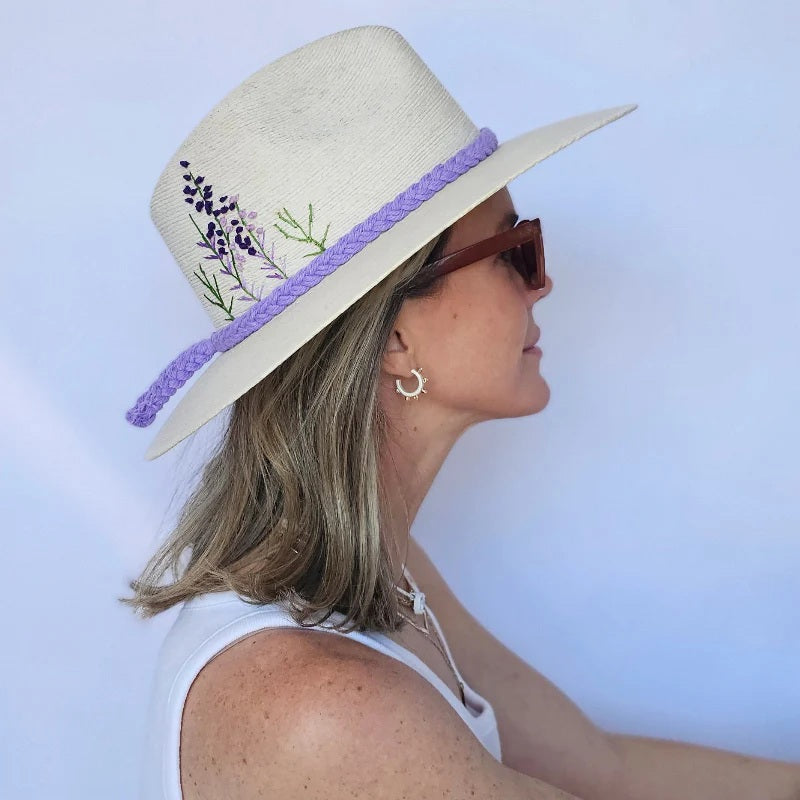 A woman in a white tank top and sunglasses poses in profile against a plain light background, wearing the BALDIZ Eggshell Palm Hat with Lavender Flower—a cream wide-brim hat with purple trim and lavender flower embroidery.