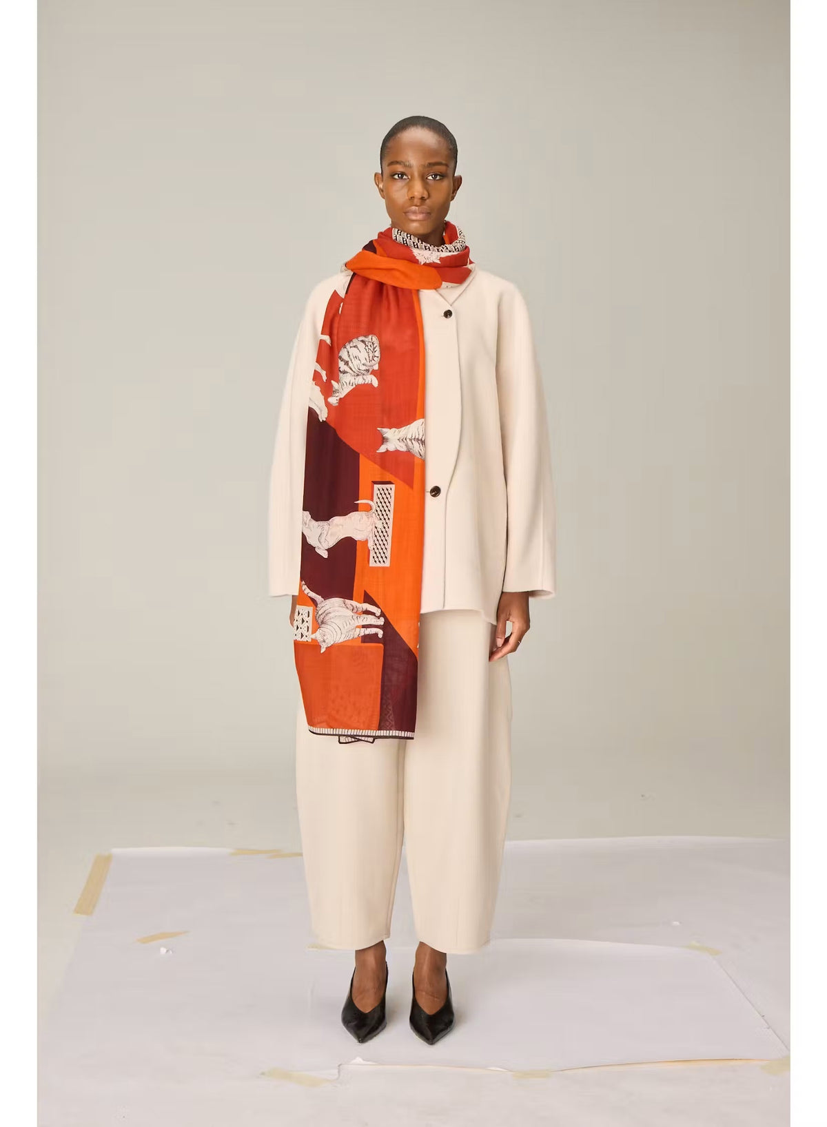 A person dressed in a cream outfit models the INOUI EDITIONS - LES CHERIS WOOL SCARF IN RED by INOUI—a 100% wool accessory made in India, featuring bold orange, red, and burgundy tones adorned with large white cat illustrations and geometric patterns—set against a plain background.