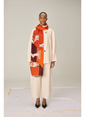 A person dressed in a cream outfit models the INOUI EDITIONS - LES CHERIS WOOL SCARF IN RED by INOUI—a 100% wool accessory made in India, featuring bold orange, red, and burgundy tones adorned with large white cat illustrations and geometric patterns—set against a plain background.