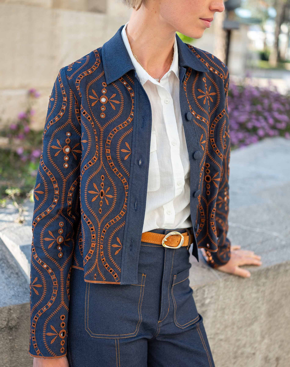 A woman stands indoors by large windows, wearing the MERSEA - FIFI JACKET—an indigo cotton jacket with cutwork embroidery—over a white shirt, paired with belted high-waisted blue pants and brown sandals. Sunlight creates grid-like shadows on the floor and wall.