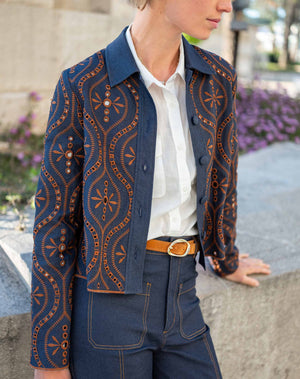 A woman stands indoors by large windows, wearing the MERSEA - FIFI JACKET—an indigo cotton jacket with cutwork embroidery—over a white shirt, paired with belted high-waisted blue pants and brown sandals. Sunlight creates grid-like shadows on the floor and wall.