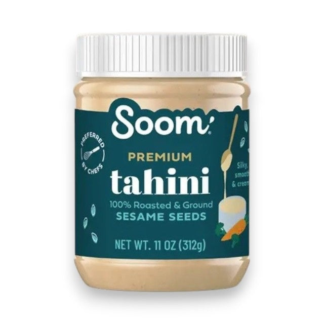 A jar of SOOM FOODS SOOM - PREMIUM TAHINI, made from 100% roasted sesame seeds and ground into a silky-smooth paste. The 11 oz (312g) jar features a dark teal label with illustrations of a spoon, carrot, and chickpea, and comes with a white lid.
