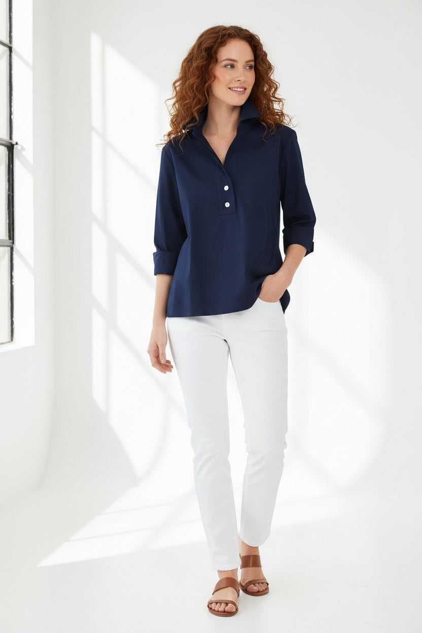 A woman with short dark hair models the HINSON WU - AILEEN 3/4 SLEEVE POLISHED COTTON SHIRT in navy, featuring a collared neckline, stretch fabric, and three buttons. She pairs it with white pants and poses against a plain light gray background.
