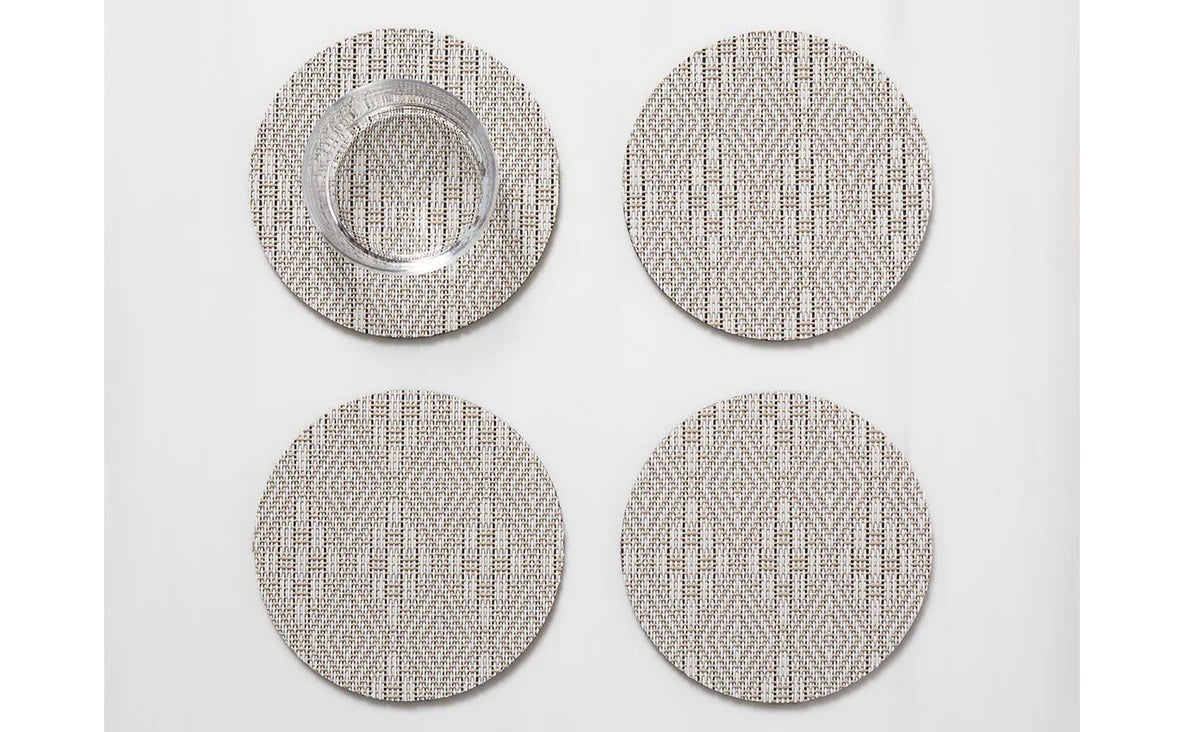 The CHILEWICH Boxwood Round Coasters Set of 4 features woven beige and white coasters with a subtle geometric pattern, combining style and function to protect your table. One holds a clear glass tumbler, all arranged on a white surface.