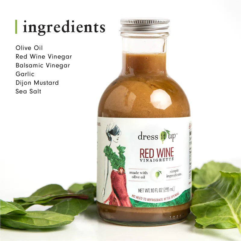 A bottle of "DRESS IT UP DRESSING - RED WINE" from the brand "DRESS IT UP DRESSING," featuring an illustrated woman in a green top and red skirt on the label. Made with olive oil and simple ingredients, it has a net weight of 10.1 fl oz (295 ml).