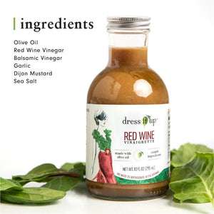 A bottle of "DRESS IT UP DRESSING - RED WINE" from the brand "DRESS IT UP DRESSING," featuring an illustrated woman in a green top and red skirt on the label. Made with olive oil and simple ingredients, it has a net weight of 10.1 fl oz (295 ml).