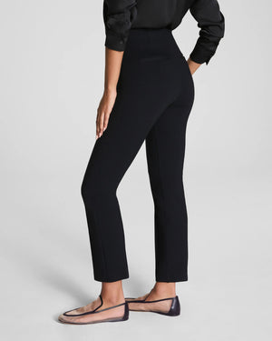 A person stands facing away in SPANX - PONTE ANKLE SLIM PANT IN BLACK by SPANX, paired with a black long-sleeve shirt and sheer pointed-toe flats. The look is simple and professional against a plain, light background.