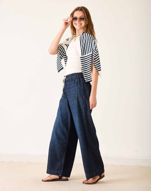 Wearing the MERSEA Londonberry Drawstring Jeans in Night Sky—a pair of wide-leg dark denim jeans with a comfortable drawstring waist—from MERSEA, styled with a cream sleeveless top, a striped shirt draped over the shoulders, and brown sandals, standing against a plain light background.