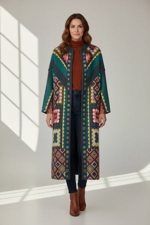 A woman stands indoors wearing the FRANCO FERRARI - REVERSIBLE WOOL COAT, its long, colorful pattern draped over a brown turtleneck, dark jeans, and brown ankle boots. Sunlight streams through a window, casting shadows on the floor and wall.