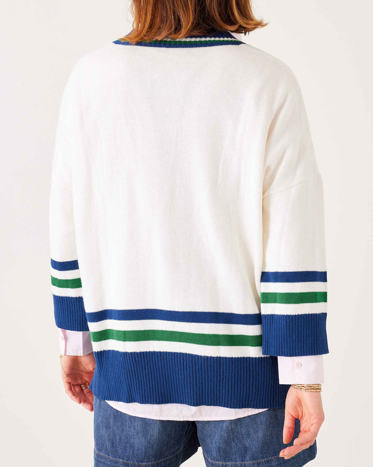 A woman wears the MERSEA - MONTAUK V-NECK SWEATER by MERSEA, a white oversized soft knit with blue and green striped collar, cuffs, and hem, ribbed v-neckline, styled over denim shorts and a light pink shirt.