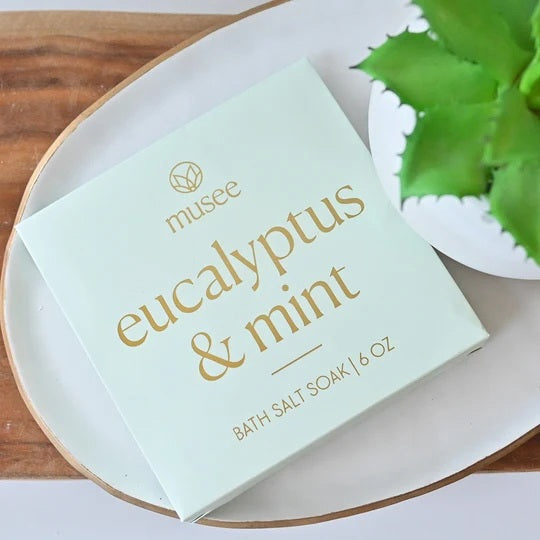 A square package labeled “MUSEE - EUCALYPTUS & MINT MINI BATH SALT SOAK” rests on a tray beside a green succulent, inviting you to enjoy the refreshing aroma of eucalyptus and mint with natural bath products from MUSEE.
