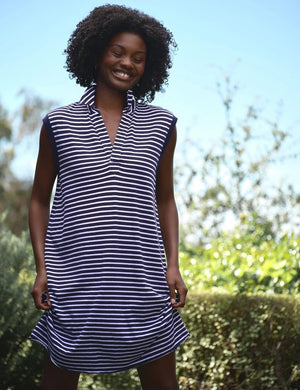 A woman stands outdoors wearing the FRANK & EILEEN MINI LILY POPOVER HENLEY TANK DRESS in Vintage Jersey Yacht Stripe. Sunlight filters through surrounding greenery, casting a soft glow on her navy and white striped, sleeveless dress from the California Heritage Collection.