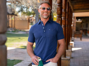 A smiling man in sunglasses stands outside by a wooden building, holding a drink and wearing the CRIQUET Top-Shelf Players Shirt in Navy. Sunlight and greenery are visible in the background.