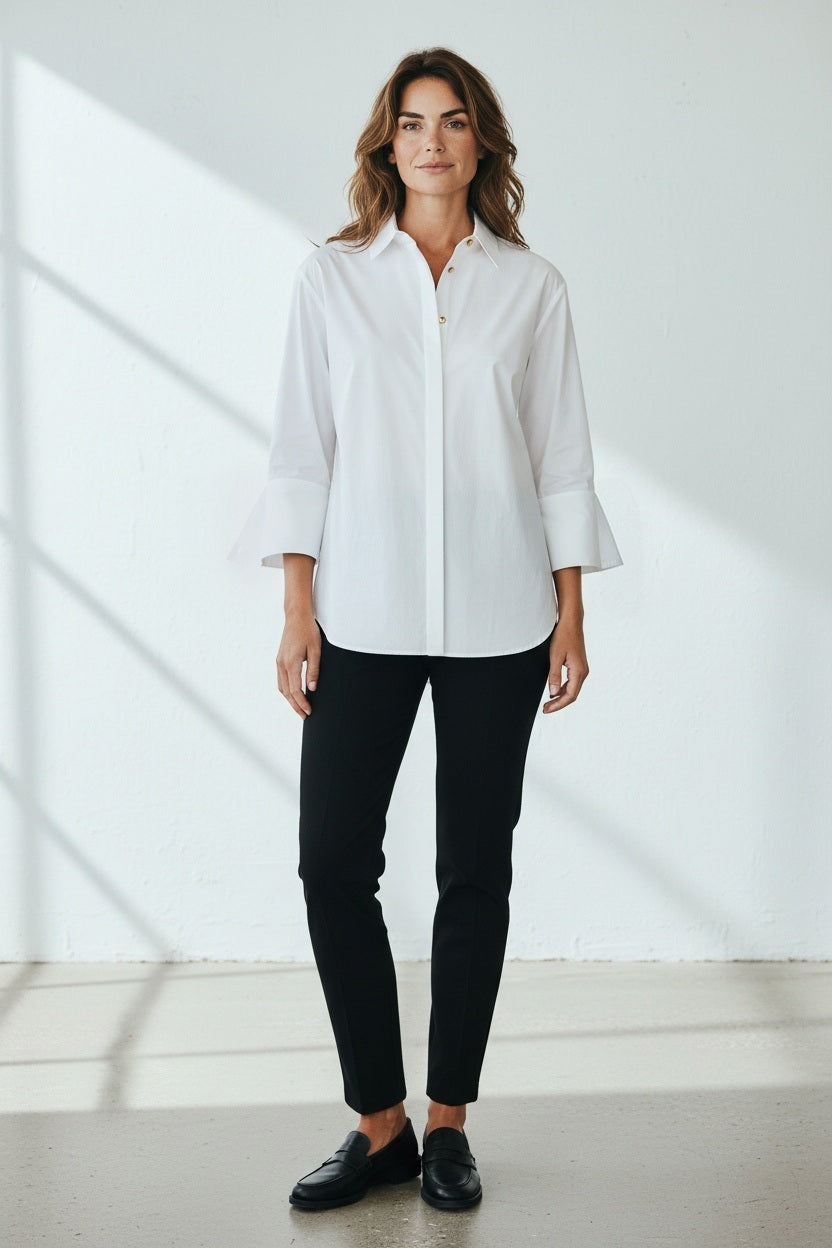 Standing against a light-colored wall with intricate molding, a woman with long hair reminiscent of Xena is turned away from the camera, showcasing the HINSON WU - XENA ZIP BACK SHIRT in navy blue. This A-line shaped top features an eye-catching gold tone zipper that extends down her back, perfectly complemented by elegant black pants.