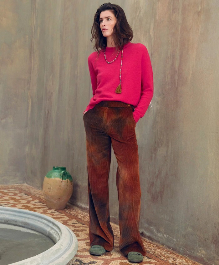 A person with long dark hair stands against a muted indoor wall, wearing a bright pink sweater, MOMONI - ISMAELTROUSER tie-dyed corduroy pants, and pink shoes. They have their hands in the patch pockets and accessorize with two long necklaces. An urn and a small water pool are placed beside them.